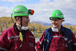 This image shows two men outdoors wearing mining or industrial work clothes, seemingly engrossed in conversation. Both men are wearing burgundy work shirts with grey reflective stripes and bright green safety helmets with reflective stickers and the Cyrillic lettering ‘БЕЗОПАСНЫЙ ТРУД’ (Safe Work). The man on the left also has a headlamp attached to his helmet and a yellow control device or measuring device hanging from his chest. Both have white respirator masks hanging loosely around their necks and are wearing safety goggles tucked into their breast pockets. The man on the right is also wearing a dark blue vest over his shirt and is looking at his colleague with a hint of a smile. In the background, an autumnal landscape stretches out with yellow-coloured trees and rolling hills under a slightly cloudy blue sky.