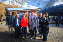 This photo shows a group of about 14 people on a joint excursion to the impressive high mountain world of Shymbulak. The group is standing on a flat gravel area in front of an imposing natural backdrop: in the background, steep, rocky mountains rise up, their peaks and flanks covered with white snow. The sun is shining brightly, creating strong contrasts and a brilliant blue sky.