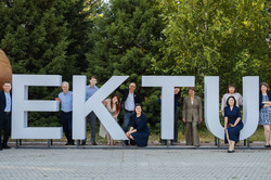 Mehrere Mitglieder der Delegation stehen und sitzen zwischen den großen weißen Buchstaben des EKTU-Schriftzugs im Außenbereich der Universität. Im Hintergrund sind Bäume zu sehen. Die Gruppe lächelt in die Kamera.