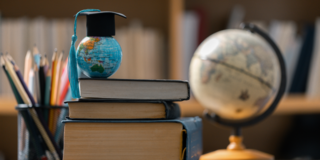 Ein kleiner Globus mit einer akademischen Doktorhut-Miniatur steht auf einem Stapel Bücher. Im Hintergrund sind ein klassischer Globus und Schreibutensilien in einer Bibliothek angedeutet.
