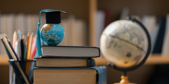 A small globe with a miniature academic mortarboard stands on a stack of books. In the background, a classic globe and writing utensils are hinted at in a library.