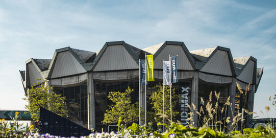 Die AudiMax der RUB steht vor einem blauen Himmel mit blühenden Blumen im Vordergrund.