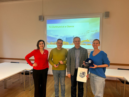 Four people are standing in a seminar room in front of a presentation slide titled “TU Dortmund at a Glance” and looking into the camera. One person is holding an award and a gift bag. The photo was taken during an official visit or exchange.