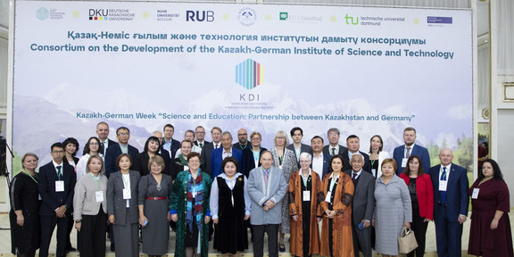 Group photo in front of a large poster with the words ‘Consortium on the Development of the Kazakh-German Institute of Science and Technology’
