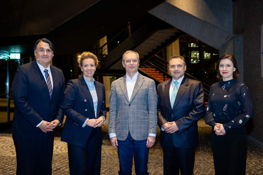 Five people stand side by side in a modern foyer with a staircase in the background and look at the camera. The group is dressed formally and poses for a group photo at an official event.
