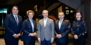 Five people stand side by side in a modern foyer with a staircase in the background and look at the camera. The group is dressed formally and poses for a group photo at an official event.