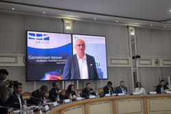 This image shows a formal conference or panel discussion taking place in a magnificent hall. It appears to be a hybrid event, with participants attending both in person and digitally.