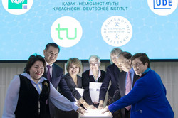 Dieses Bild zeigt eine feierliche Zeremonie, die den Start oder die Einweihung einer Kooperation markiert. Im Zentrum stehen sieben Personen (fünf Frauen und zwei Männer), die sich um ein kleines, weißes Stehpult gruppiert haben. Alle Beteiligten haben ihre Hände gleichzeitig auf eine leuchtende Glasplatte auf dem Pult gelegt, was wie ein symbolischer „Startknopf“ wirkt.