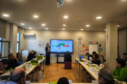 Teilnehmende sitzen in einem Workshopraum an Tischen und arbeiten mit Unterlagen und Laptops, während eine Referentin vorne den Ablauf des Programms im Rahmen eines Workshops präsentiert.