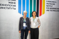 This image shows two women standing smiling in front of a decoratively designed wall in a bright interior. In the centre of the wall is a large, modern logo consisting of seven vertical colour bars of varying lengths, which together form a hexagonal shape; the colours range from shades of blue and green on the left to black, red and yellow on the right. The logo is framed by two blocks of text: on the left is the Kazakh name of the institute in Cyrillic script and on the right is the German equivalent, ‘KASACHISCH - DEUTSCHES INSTITUT FÜR WISSENSCHAFT UND TECHNOLOGIE’ (Kazakh-German Institute for Science and Technology).