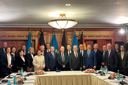 Eine große Gruppe von Vertreter*innen steht für ein gemeinsames Foto in einem repräsentativen Konferenzraum. Im Hintergrund sind deutsche und kasachische Flaggen sowie ein Wappen zu sehen. Vor der Gruppe steht ein großer Konferenztisch mit Mikrofonen und Blumenarrangements.