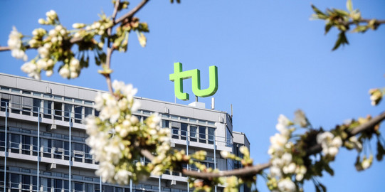 Gebäude der Technischen Universität Dortmund mit dem grünen ‚tu‘-Logo auf dem Dach. Im Vordergrund blühende Äste vor blauem Himmel.