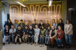 Group photo of international alumni of Studienbrücke during a tour of the BRYCK Startup Alliance. Around 20 people are sitting and standing in front of a large, illuminated ‘BRYCK’ sign in a modern interior.