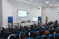 Ein Hörsaal der East Kazakhstan Technical University (EKTU) mit zahlreichen Teilnehmenden. Vorne sitzen drei Referentinnen an einem Podium, hinter ihnen sind Präsentationsfolien auf einer Leinwand und einem Bildschirm zu sehen. Banner der University Alliance Ruhr und der EKTU flankieren die Bühne, während das Publikum den Vorträgen aufmerksam folgt.