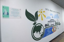 Dieses Bild zeigt eine hell gestaltete Wand in einem Innenraum, die als Informations- und Motivationsfläche dient. Auf der linken Seite ist eine transparente Acryltafel mit silbernen Metallstiften an der Wand befestigt, die ein grünliches Porträt des Gelehrten Alexander von Humboldt sowie den dreisprachigen Schriftzug „Alexander von Humboldt Interdisciplinary Centre for Sustainable Development“ trägt. Rechts daneben erstreckt sich ein großflächiges Wandbild in einem modernen, flachen Grafikstil unter der Überschrift „EKTU - Green Leader of Kazakhstan“. Die Illustration zeigt eine stilisierte Weltkugel, aus der zwei große, dunkelgrüne Blätter sprießen, überragt von einer strahlend gelben Sonne mit kräftigen Strahlen. Im rechten Teil des Wandbildes sind Symbole für Nachhaltigkeit und Fortschritt kombiniert: die Silhouette eines klassischen Universitätsgebäudes mit einer Flagge, Windkrafträder, Solarpanele, eine Person auf einem Fahrrad sowie ein gewundener blauer Flussweg. Am unteren Rand des Bildes ist zudem eine Reihe bunter, hexagonaler Icons zu sehen, welche die globalen Nachhaltigkeitsziele (SDGs) repräsentieren. Die gesamte Komposition wirkt durch die Nutzung von viel Weißraum sowie den Farben Grün, Gelb und Blau sehr frisch, zukunftsorientiert und akademisch.