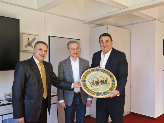 Drei Männer stehen in einem Büro nebeneinander und halten gemeinsam einen dekorativen Teller mit einer Gebäudeabbildung. Die Aufnahme zeigt eine symbolische Geschenkübergabe im Rahmen eines offiziellen Treffens.