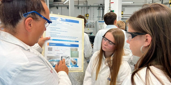 Wissenschaftlerin zeigt zwei Schulerinnen im Labor in Laborkitteln eine Tabelle im Labor.