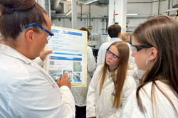 Wissenschaftlerin zeigt zwei Schulerinnen im Labor in Laborkitteln eine Tabelle im Labor.